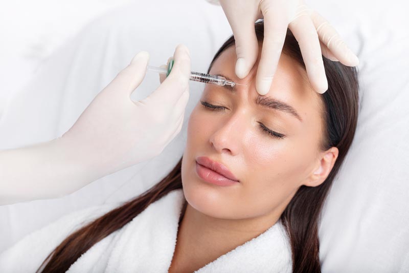 behandlungen-botox Dr. Sarah Ophoff bei der ästhetischen Gesichtsbehandlung in ihrer Praxis am Tegernsee – professionell, einfühlsam und präzise. Sie trägt einen weißen Kittel mit Namensschild. Ihre Haare sind in einem Pferdeschwanze gebunden. Botox Behandlung Zornesfalte