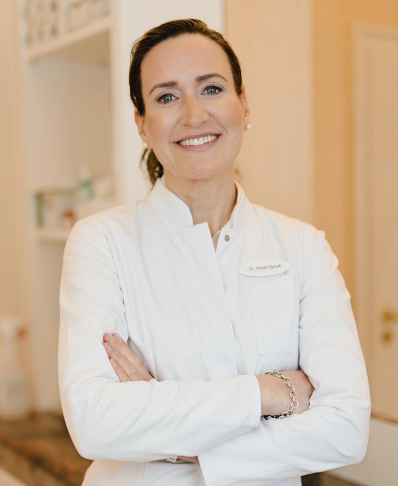dr-sarah-ophoff Dr. Sarah Ophoff bei der ästhetischen Gesichtsbehandlung in ihrer Praxis am Tegernsee – professionell, einfühlsam und präzise. Sie trägt einen weißen Kittel mit Namensschild. Ihre Haare sind in einem Pferdeschwanze gebunden.